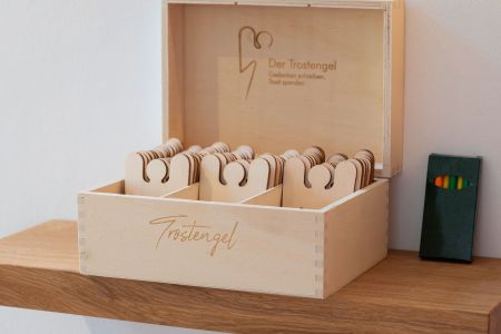 Trauerforum Leimbachtal Pietät Siegen – Box Trostengel aus Holz auf einem Regal mit bunten Stiften